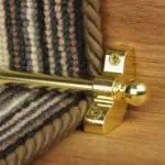 Jubilee Stair Carpet Runner Rod in shiny brass installed on wooden stairs with a striped runner